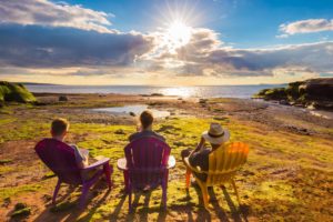 Three people seated in chairs by coast watching sunset