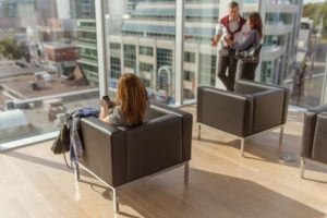 Woman seated in chair looking at mobile phone facing toward windows of office building
